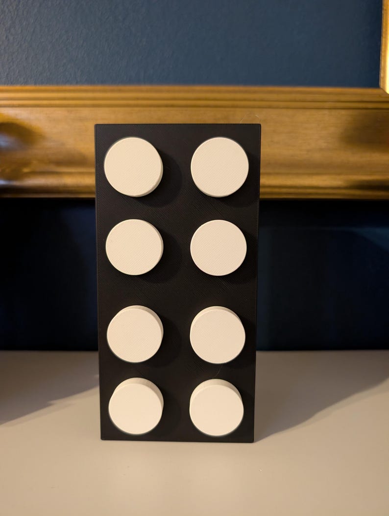 May include: A black rectangular decorative object with eight white circular accents arranged in two columns of four. The object is standing upright on a white surface, with a dark blue wall and gold-framed artwork in the background.