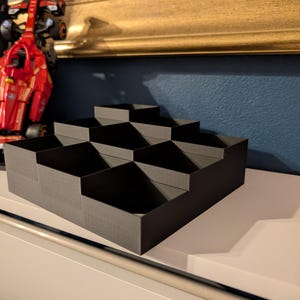 May include: A black, stepped organizer with multiple compartments. The organizer is made of a matte material and is designed to hold small items. The design is geometric and modern, suitable for desk or shelf organization.
