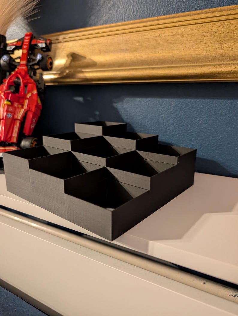 May include: A black, stepped organizer with multiple compartments. The organizer is made of a matte material and is designed to hold small items. The design is geometric and modern, suitable for desk or shelf organization.