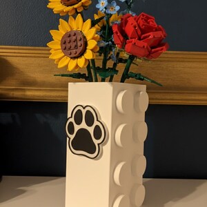 May include: A white vase with a black paw print sticker, holding a bouquet of yellow sunflowers, red roses, and blue flowers. The vase has a brick-like design with protruding circular elements.