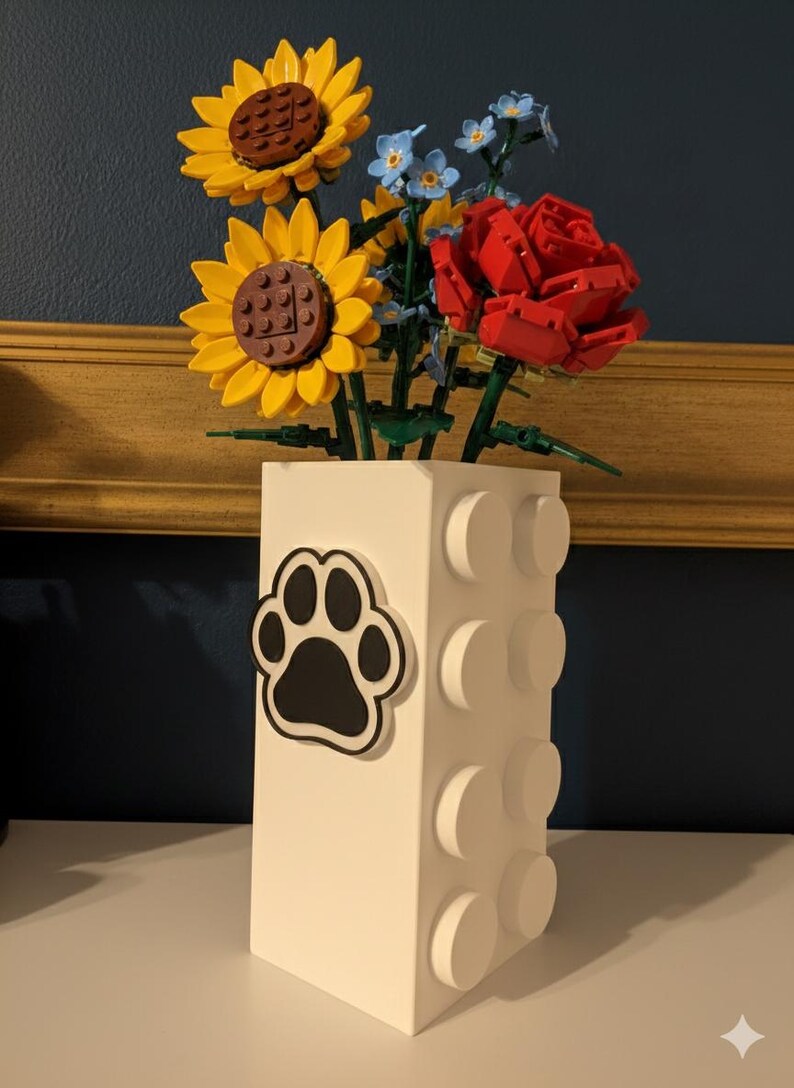 May include: A white vase with a black paw print sticker, holding a bouquet of yellow sunflowers, red roses, and blue flowers. The vase has a brick-like design with protruding circular elements.