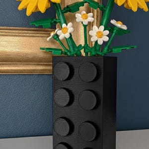 May include: A black vase with a textured surface, resembling a large building block, holds a bouquet of artificial flowers. The bouquet includes two large sunflowers with yellow petals and brown centers, along with smaller white daisies.