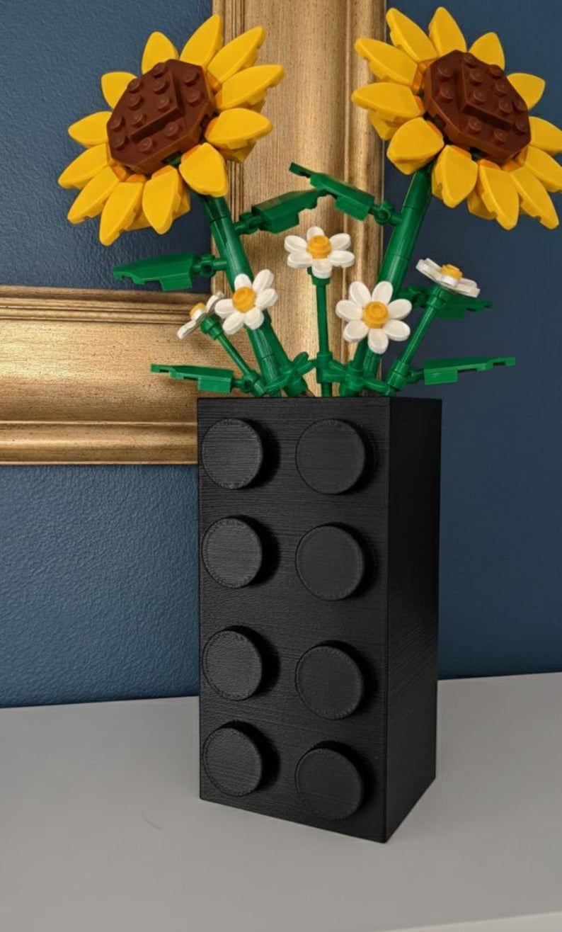 May include: A black vase with a textured surface, resembling a large building block, holds a bouquet of artificial flowers. The bouquet includes two large sunflowers with yellow petals and brown centers, along with smaller white daisies.