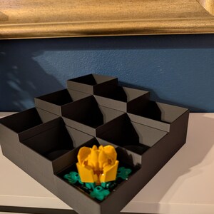 May include: A dark gray, tiered organizer with multiple compartments. A yellow and green flower is placed in one of the compartments. The organizer is on a white surface, with a gold-framed picture in the background.