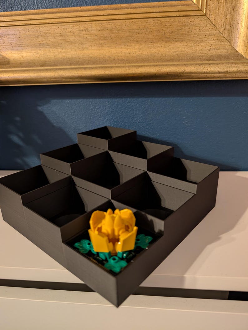 May include: A dark gray, tiered organizer with multiple compartments. A yellow and green flower is placed in one of the compartments. The organizer is on a white surface, with a gold-framed picture in the background.
