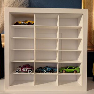 May include: A white, multi-compartment display case holding toy cars. The case has a grid of small cubicles, with several toy cars in various colors, including yellow, green, blue, and maroon. The cars are miniature replicas of different vehicle models.