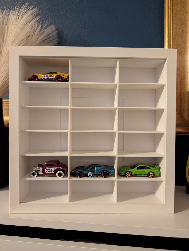 May include: A white, multi-compartment display case holding toy cars. The case has a grid of small cubicles, with several toy cars in various colors, including yellow, green, blue, and maroon. The cars are miniature replicas of different vehicle models.