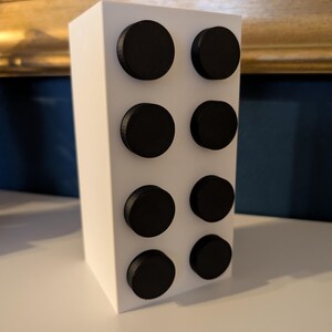 May include: A white rectangular block with eight evenly spaced, black circular accents. The object is positioned on a white surface, with a gold-framed artwork and a blue wall in the background.
