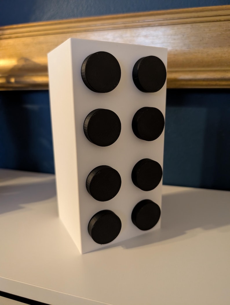 May include: A white rectangular block with eight evenly spaced, black circular accents. The object is positioned on a white surface, with a gold-framed artwork and a blue wall in the background.