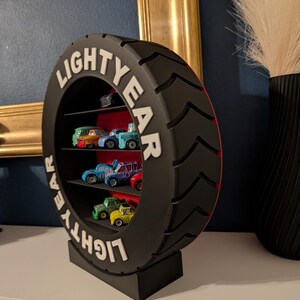 May include: A black, tire-shaped shelf unit with the word "LIGHTYEAR" in white lettering. The shelf contains several toy cars of various colors. The unit is on a white surface, with a dark blue wall in the background.