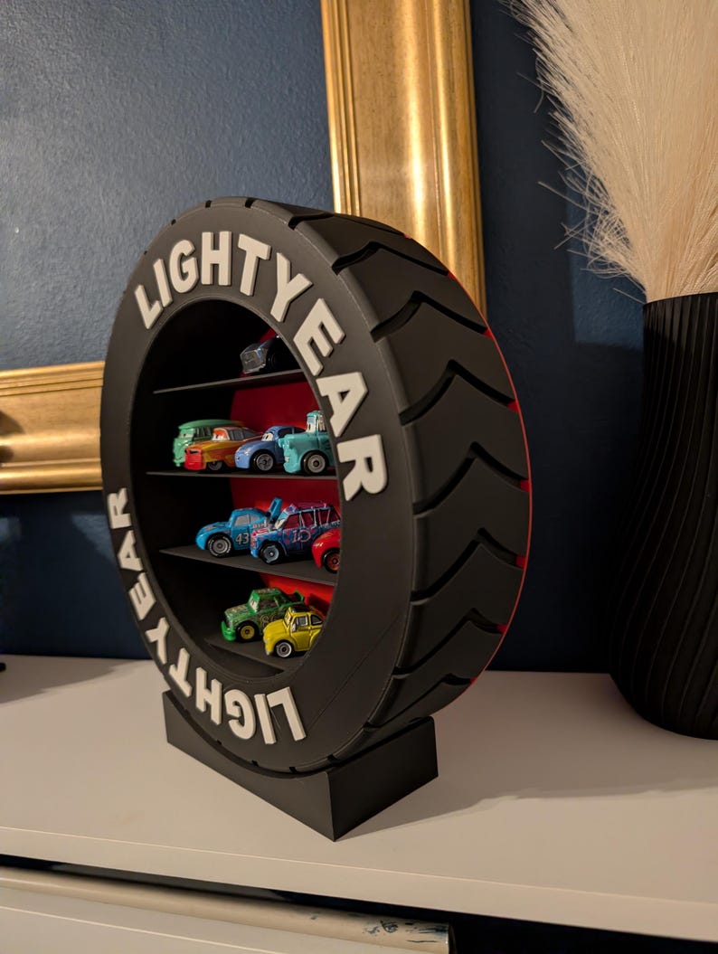 May include: A black, tire-shaped shelf unit with the word "LIGHTYEAR" in white lettering. The shelf contains several toy cars of various colors. The unit is on a white surface, with a dark blue wall in the background.