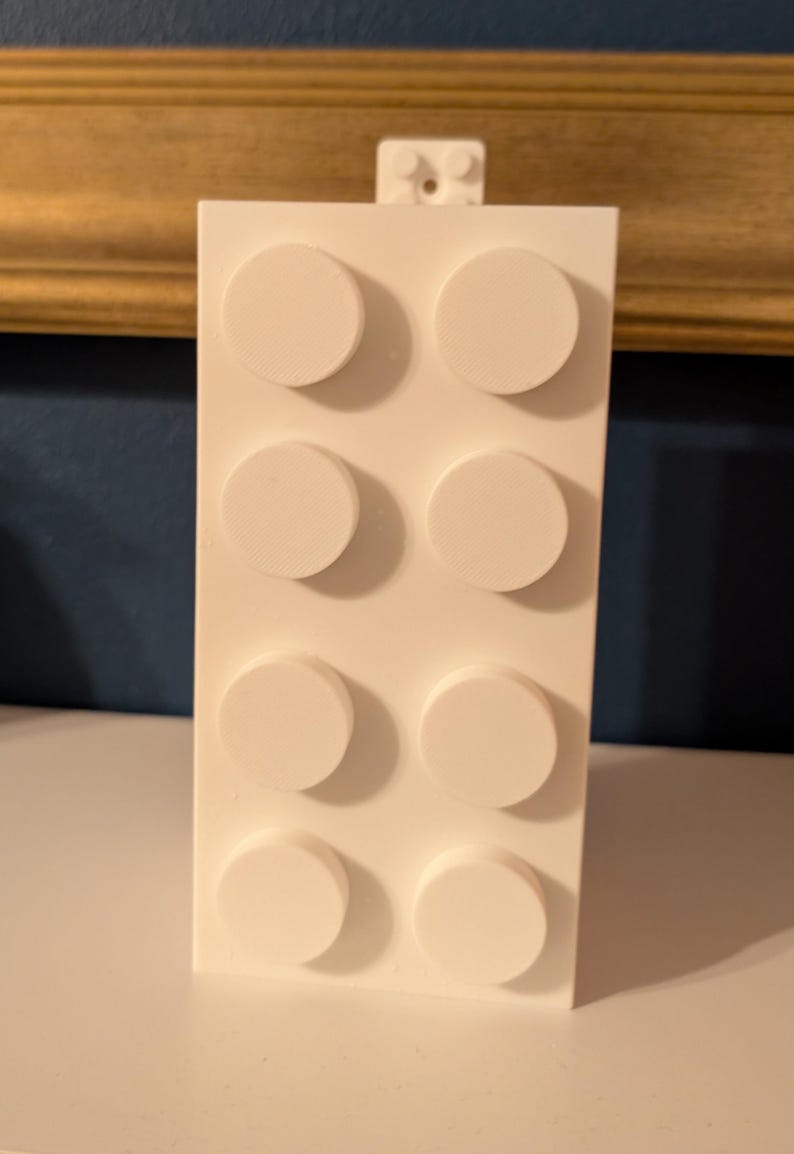 Op de afbeelding: Een witte bouwsteenspeelgoed met acht verhoogde cirkelvormige noppen aan de voorkant. Het speelgoed heeft een rechthoekige vorm met een kleiner verbindingsstuk aan de bovenkant. De achtergrond omvat een goudomrand kader en een donkerblauwe muur.