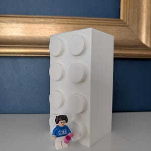 May include: A white, rectangular block-shaped decorative item with six raised circular studs. A small Lego figure in a blue sweater and white pants is standing in front of the block, holding a pink object. The background includes a blue wall and a gold-framed picture.
