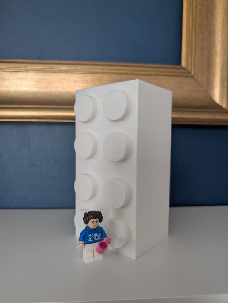 May include: A white, rectangular block-shaped decorative item with six raised circular studs. A small Lego figure in a blue sweater and white pants is standing in front of the block, holding a pink object. The background includes a blue wall and a gold-framed picture.