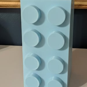 Op de afbeelding: Een lichtblauwe, rechthoekige blokvormige container met acht cirkelvormige noppen aan de voorkant. Een kleiner blok met vier noppen is aan de bovenkant bevestigd. De container is waarschijnlijk een pennenhouder of bureau-organizer.