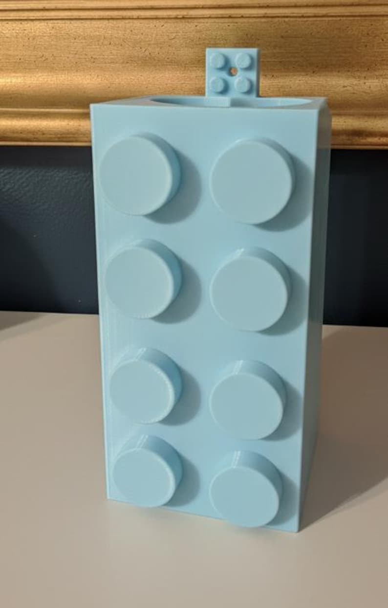 Op de afbeelding: Een lichtblauwe, rechthoekige blokvormige container met acht cirkelvormige noppen aan de voorkant. Een kleiner blok met vier noppen is aan de bovenkant bevestigd. De container is waarschijnlijk een pennenhouder of bureau-organizer.