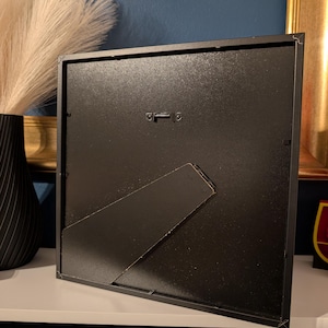 May include: A black square picture frame with a built-in easel back. The frame is made of metal and has a matte finish. The back of the frame has a metal hanger for wall mounting. The frame is sitting on a white surface.