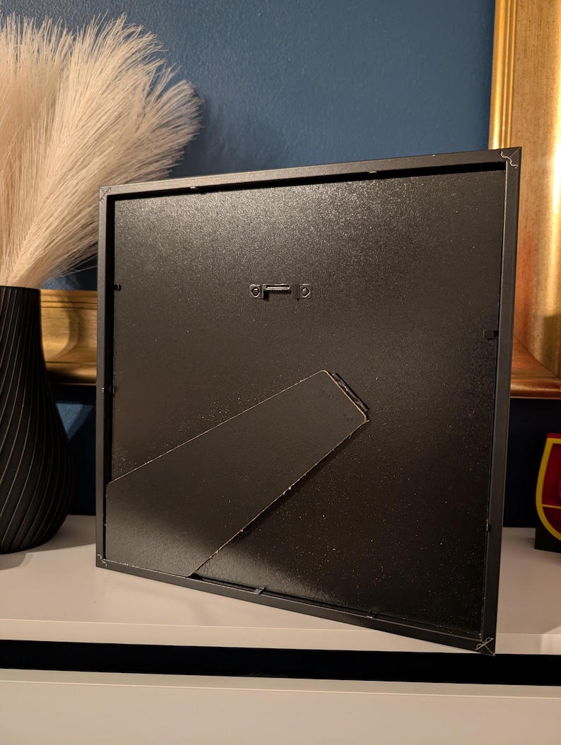 May include: A black square picture frame with a built-in easel back. The frame is made of metal and has a matte finish. The back of the frame has a metal hanger for wall mounting. The frame is sitting on a white surface.