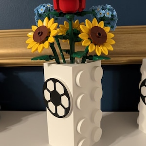 May include: A white vase with a soccer ball design, holding a bouquet of Lego flowers. The bouquet includes a red rose, yellow sunflowers, and blue forget-me-nots. The vase is made of white Lego bricks.