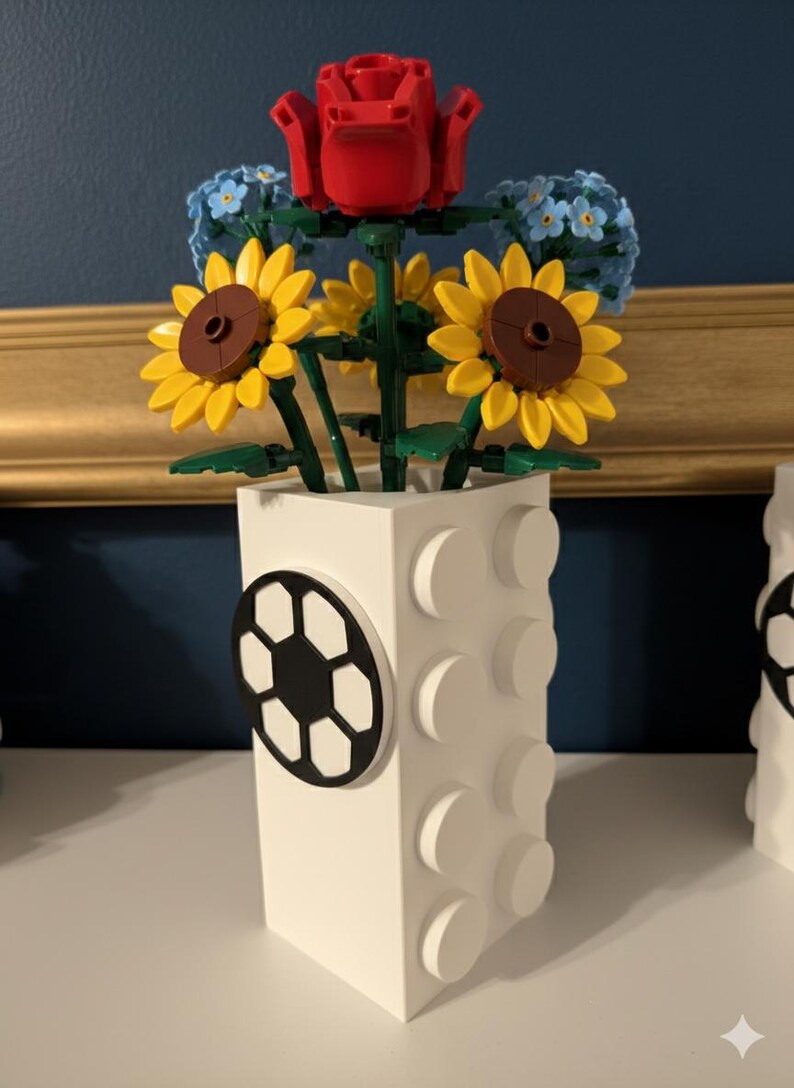May include: A white vase with a soccer ball design, holding a bouquet of Lego flowers. The bouquet includes a red rose, yellow sunflowers, and blue forget-me-nots. The vase is made of white Lego bricks.