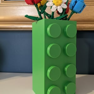 May include: A green vase shaped like a building block, holding colorful flowers made of toy blocks. The flowers include red tulips, yellow tulips, white daisies, and blue tulips. The vase has eight circular studs on the front.