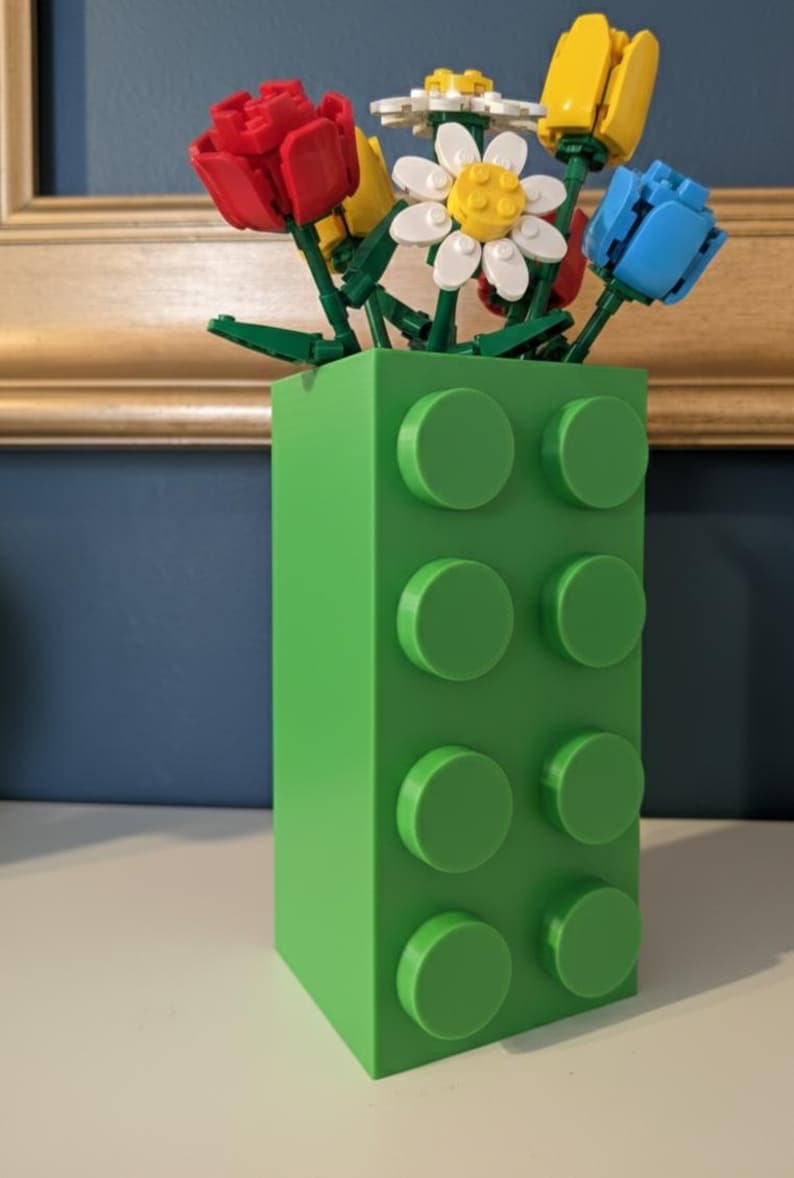 May include: A green vase shaped like a building block, holding colorful flowers made of toy blocks. The flowers include red tulips, yellow tulips, white daisies, and blue tulips. The vase has eight circular studs on the front.