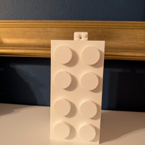 Op de afbeelding: Een witte bouwsteen met acht verhoogde cirkelvormige noppen. De blok is rechthoekig en heeft een klein verbindingsstuk aan de bovenkant. De achtergrond heeft een donkerblauwe muur en een goudkleurig frame.