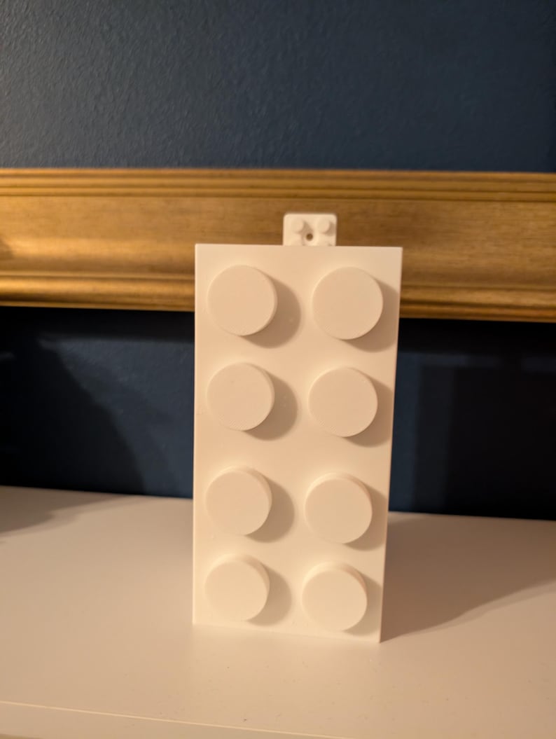 Op de afbeelding: Een witte bouwsteen met acht verhoogde cirkelvormige noppen. De blok is rechthoekig en heeft een klein verbindingsstuk aan de bovenkant. De achtergrond heeft een donkerblauwe muur en een goudkleurig frame.