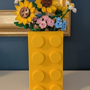 May include: A bright yellow vase made of interlocking blocks holds a colorful arrangement of flowers. The flowers include yellow sunflowers, pink and blue blossoms, and white accents. The vase has a rectangular shape with eight circular studs.