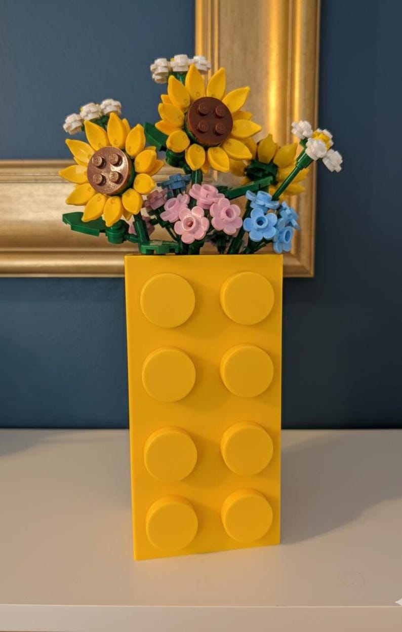 May include: A bright yellow vase made of interlocking blocks holds a colorful arrangement of flowers. The flowers include yellow sunflowers, pink and blue blossoms, and white accents. The vase has a rectangular shape with eight circular studs.