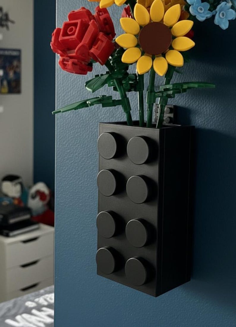 Op de afbeelding: Een zwarte, aan de muur bevestigde vaas in de vorm van een grote bouwsteen, met kleurrijke Lego-bloemen. De bloemen zijn onder meer een rode roos, een gele zonnebloem en blauwe bloesems. De vaas heeft acht ronde noppen.