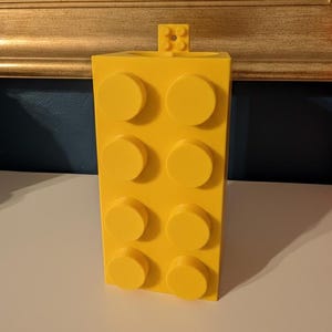 Op de afbeelding: Een felgele bouwsteen-vormige container met acht verhoogde cirkelvormige noppen aan de voorkant. Een kleiner blok is aan de bovenkant bevestigd. De container is gemaakt van plastic en staat op een wit oppervlak.