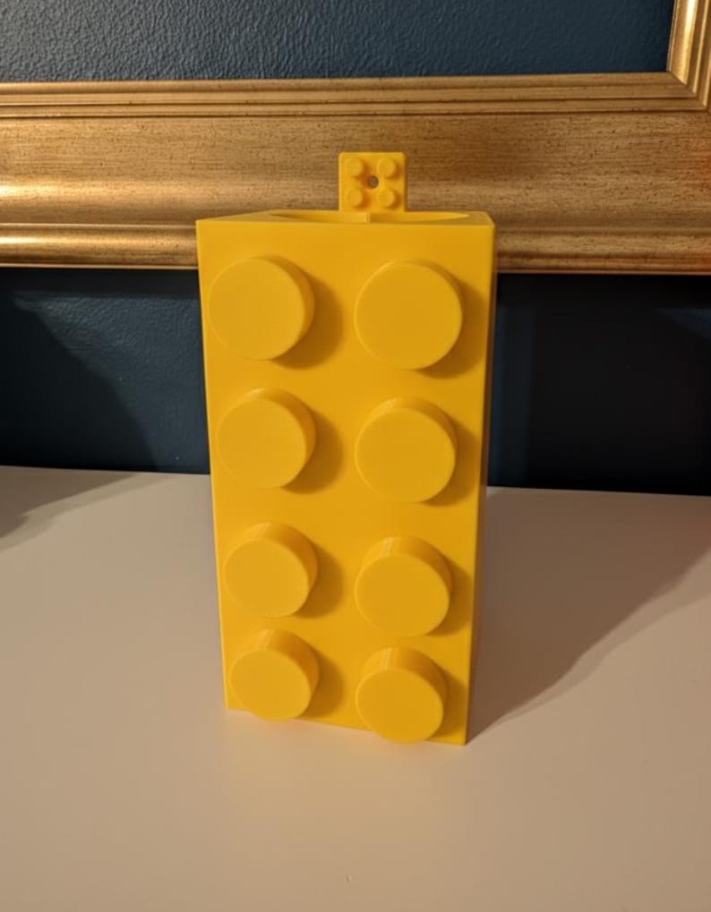 Op de afbeelding: Een felgele bouwsteen-vormige container met acht verhoogde cirkelvormige noppen aan de voorkant. Een kleiner blok is aan de bovenkant bevestigd. De container is gemaakt van plastic en staat op een wit oppervlak.