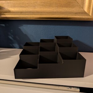May include: A black, tiered organizer with multiple compartments. The organizer is made of a matte material and is designed to hold various items. It sits on a white surface against a blue wall, with a gold-framed picture in the background.