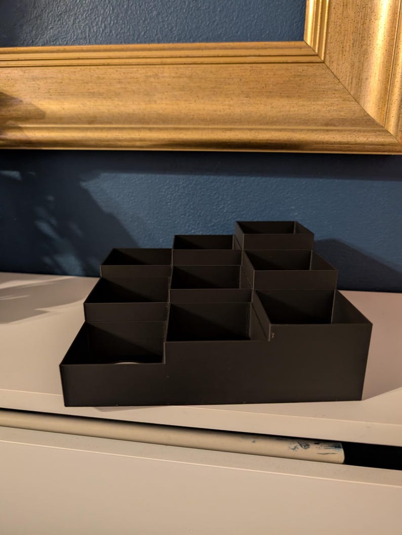 May include: A black, tiered organizer with multiple compartments. The organizer is made of a matte material and is designed to hold various items. It sits on a white surface against a blue wall, with a gold-framed picture in the background.