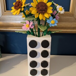 Puede incluir: Un jarr&oacute;n rectangular blanco con ocho acentos circulares negros contiene un ramo de flores coloridas. Las flores incluyen girasoles amarillos, flores rosas, azules y blancas, todas hechas de bloques de pl&aacute;stico.