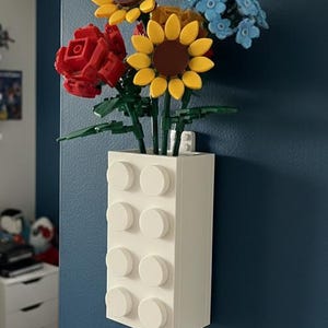 Op de afbeelding: Een witte rechthoekige vaas van in elkaar grijpende blokken bevat een boeket kleurrijke bloemen. De bloemen zijn onder meer gele zonnebloemen, een rode roos en blauwe bloesems. De vaas is aan een donkerblauwe muur bevestigd.
