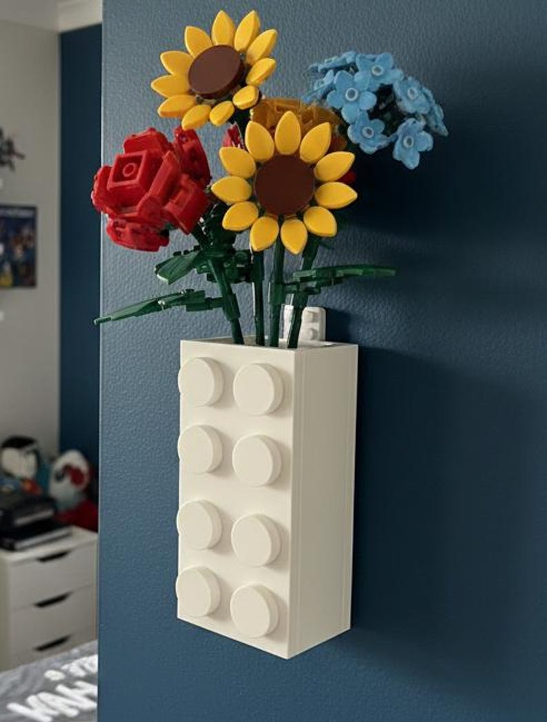 Op de afbeelding: Een witte rechthoekige vaas van in elkaar grijpende blokken bevat een boeket kleurrijke bloemen. De bloemen zijn onder meer gele zonnebloemen, een rode roos en blauwe bloesems. De vaas is aan een donkerblauwe muur bevestigd.
