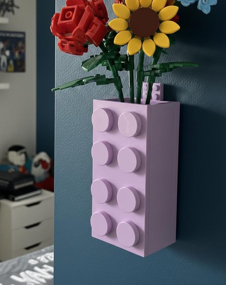 Op de afbeelding: Een lavendelkleurige vaas in de vorm van een bouwsteen, met kleurrijke kunstbloemen. De bloemen zijn onder meer een rode roos, een gele zonnebloem en blauwe bloemen. De vaas is aan een donkerblauwe muur bevestigd.