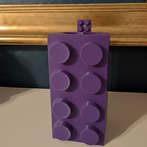 Op de afbeelding: Een paarse, rechthoekige blokvormige container met acht cirkelvormige noppen aan de voorkant en een kleiner blokje bovenop. De container is gemaakt van plastic en lijkt op een bouwsteen.