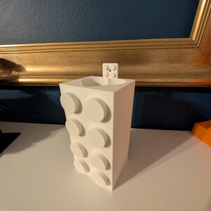 Op de afbeelding: Een witte, rechthoekige vaas met een ontwerp van verhoogde, cirkelvormige elementen. De vaas heeft een klein, rechthoekig stukje aan de bovenkant bevestigd. De vaas staat op een wit oppervlak.