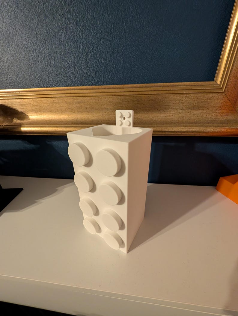 Op de afbeelding: Een witte, rechthoekige vaas met een ontwerp van verhoogde, cirkelvormige elementen. De vaas heeft een klein, rechthoekig stukje aan de bovenkant bevestigd. De vaas staat op een wit oppervlak.