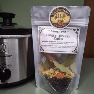 May include: A clear pouch labeled "Forest-Hearth Ember" with botanical ingredients, including orange peel, bay leaf, and juniper berries. The pouch is next to a silver slow cooker with a black dial.