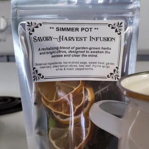 May include: A clear, sealed bag labeled "Simmer Pot Savory-Harvest Infusion" contains dried herbs and citrus slices. The label describes the blend as a revitalizing mix of garden-grown herbs and bright citrus. A white enamel mug is partially visible.
