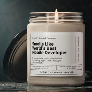 Könnte beinhalten: Eine beige Sojawachskerze in einem Glasgefäß mit schwarzem Deckel. Die Kerze ist mit "Smells Like World's Best Mobile Developer" beschriftet. Das Etikett lautet außerdem "Ich schätze Ihr Engagement für die Entwicklung von Apps, die begeistern und verbinden." und "50-60 Stunden Brenndauer 9 oz (250g)". Das Etikett lautet außerdem "Scent the mood, stay lit."