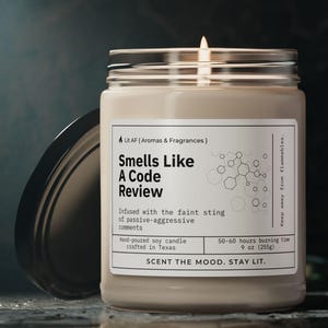 Puede incluir: Un tarro de vela de vidrio transparente con una vela de cera beige en su interior. La vela está encendida y la llama es visible. El tarro tiene una tapa negra y una etiqueta blanca que dice "Smells Like A Code Review" con el texto "Indused with the faint sting of passive-aggressive comments" y "SCENT THE MOOD. STAY LIT."
