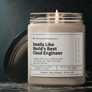 May include: A beige soy candle with a black label that reads "Smells Like World's Best Cloud Engineer". The label also includes the text "I appreciate your dedication to keeping our data flying high and secure." and "50-60 hours burning time 9 oz (250g)". The candle is lit and the flame is visible.
