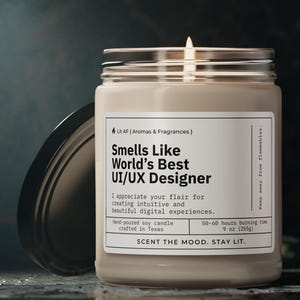 Op de afbeelding: Een beige sojawas kaars in een heldere glazen pot met een zwarte deksel. De kaars is gelabeld "Smells Like World's Best UI/UX Designer". Het label vermeldt ook "Ik waardeer je flair voor het creëren van intuïtieve en mooie digitale ervaringen." en "Handgegoten sojawas kaars gemaakt in Texas. 50-60 uur brandtijd. 9 oz (250 g). De sfeer creëren, licht aan laten."