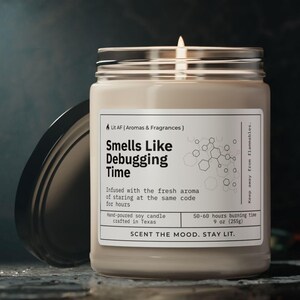 Puede incluir: Una vela de soja beige en un tarro de vidrio con tapa negra. La vela está etiquetada como "Smells Like Debugging Time" y tiene un gráfico de una estructura de código. La vela está hecha a mano y tiene un tiempo de combustión de 50-68 horas.