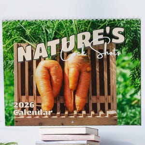 May include: A 2026 calendar featuring the text "NATURE'S Cock Shots" with two large, orange carrots in a wooden crate. The background shows green foliage.
